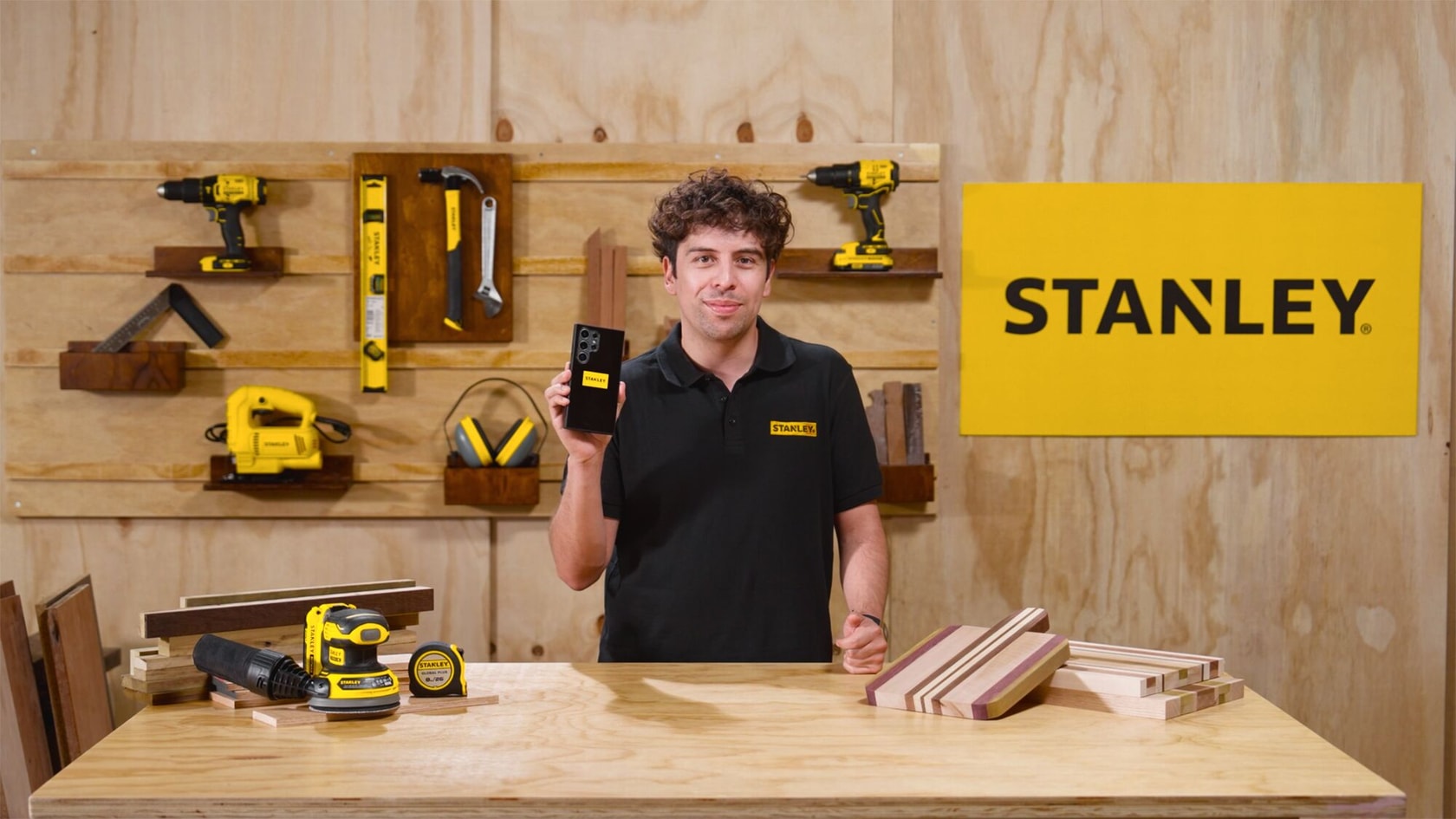 Una persona con una camisa negra de la marca STANLEY está en un taller de carpintería, sosteniendo una herramienta STANLEY. En el taller hay varias herramientas STANLEY en la pared y un cartel amarillo STANLEY. Sobre la mesa hay bloques de madera y herramientas.