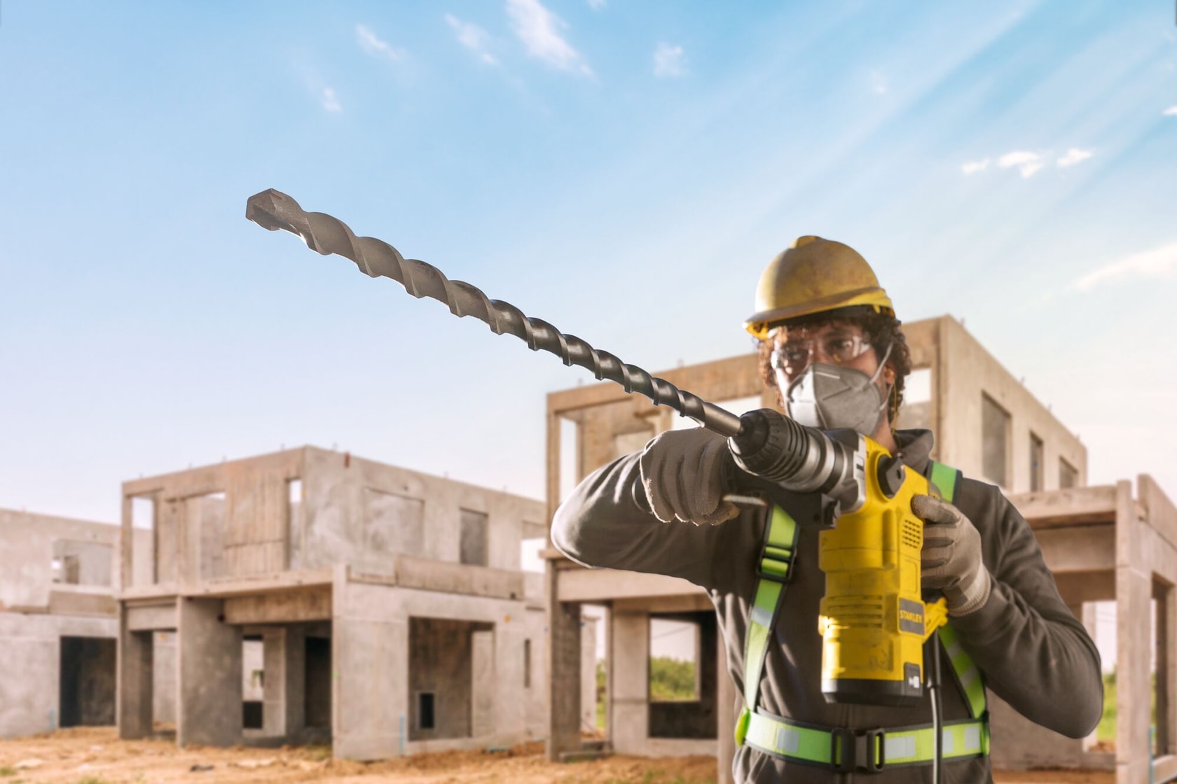 Un trabajador de la construcción con equipo de seguridad sostiene un taladro rotativo amarillo STANLEY con una broca larga. El trabajador está al aire libre en un sitio de construcción con estructuras de concreto sin terminar en el fondo.